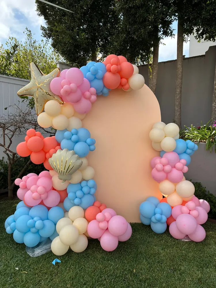 A pastel balloon arch around an event backdrop on a lawn.